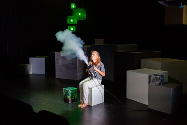 A woman sitting on a box holding a smoke machine & blowing out while the smoke goes up - there are 3 green lights hanging in the background.     Photo Credit Orange Schmorange